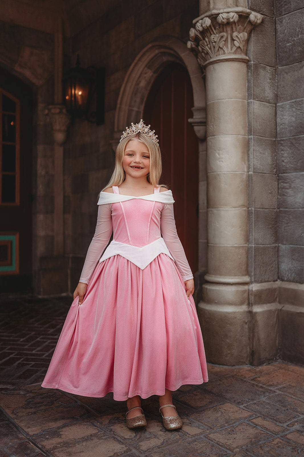 A young girl in an exquisite Portrait Collection Sleeping Beauty gown poses in a castle setting. The pink stretch velvet gown has an off the shoulder neckline with two straps. The long sleeves are light pink mesh. The dress has sewn in tulle and a cotton liner to add fullness in the skirt.