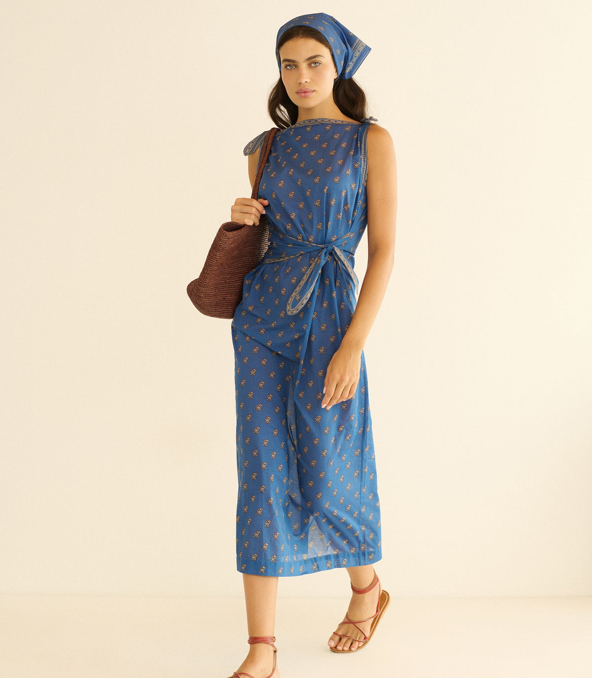 Woman wearing a blue paisley dress, holding a brown bag on a beige background