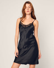 A woman with long brown hair wears a luxurious Womens Silk Lace Slip Dress in Black by Petite Plume, showcasing delicate lace at the neckline, set against a plain background.
