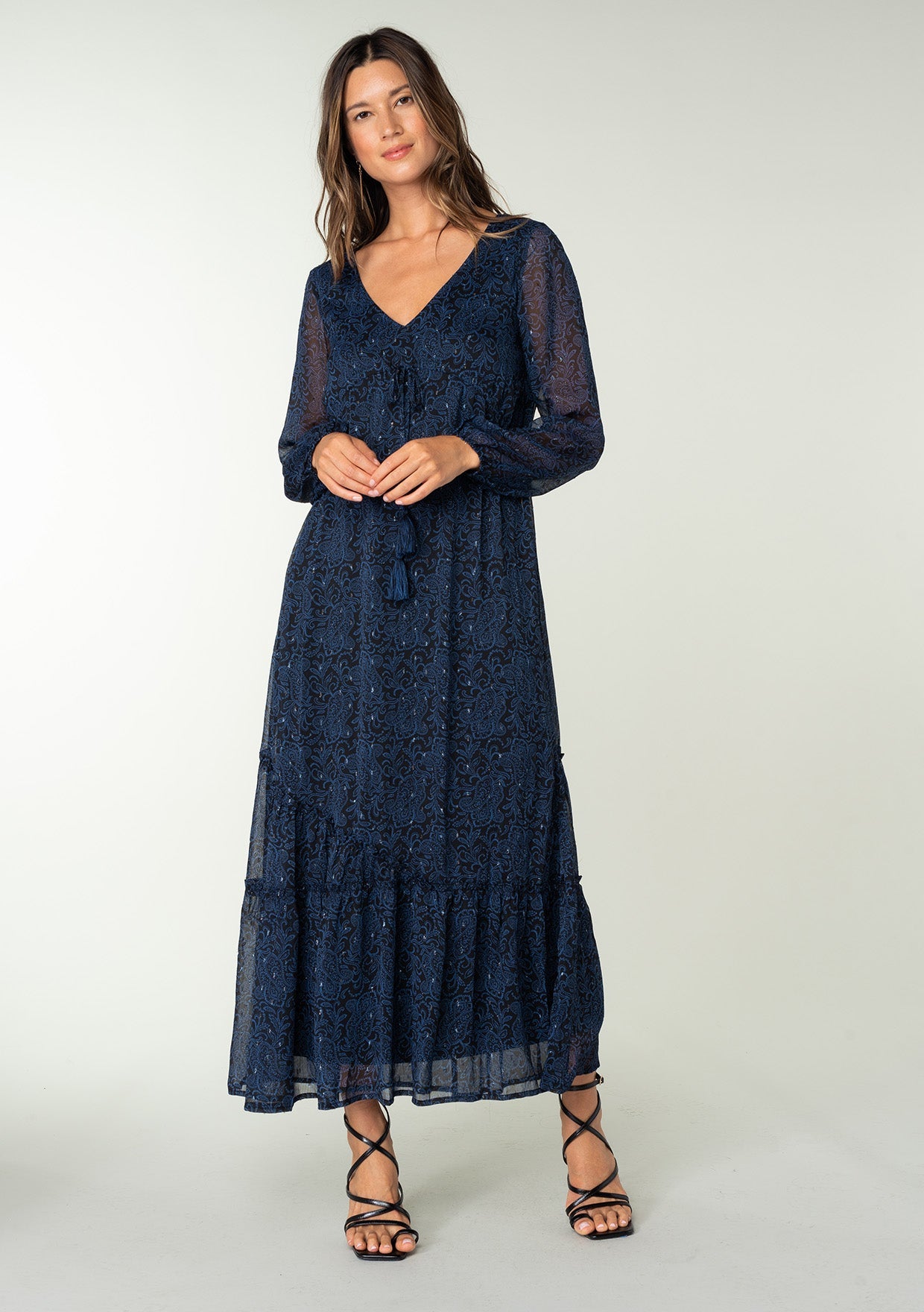 [Color: Black/Blue] A full body front facing image of a brunette model wearing a best selling navy blue chiffon maxi dress with black paisley print and light catching metallic clip dot details. Perfect for special occasions, with sheer long sleeves, a v neckline, an empire waist with drawstring tassel tie detail, and an asymmetric ruffled trimmed tiered skirt.