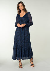 [Color: Black/Blue] A full body front facing image of a brunette model wearing a best selling navy blue chiffon maxi dress with black paisley print and light catching metallic clip dot details. Perfect for special occasions, with sheer long sleeves, a v neckline, an empire waist with drawstring tassel tie detail, and an asymmetric ruffled trimmed tiered skirt.