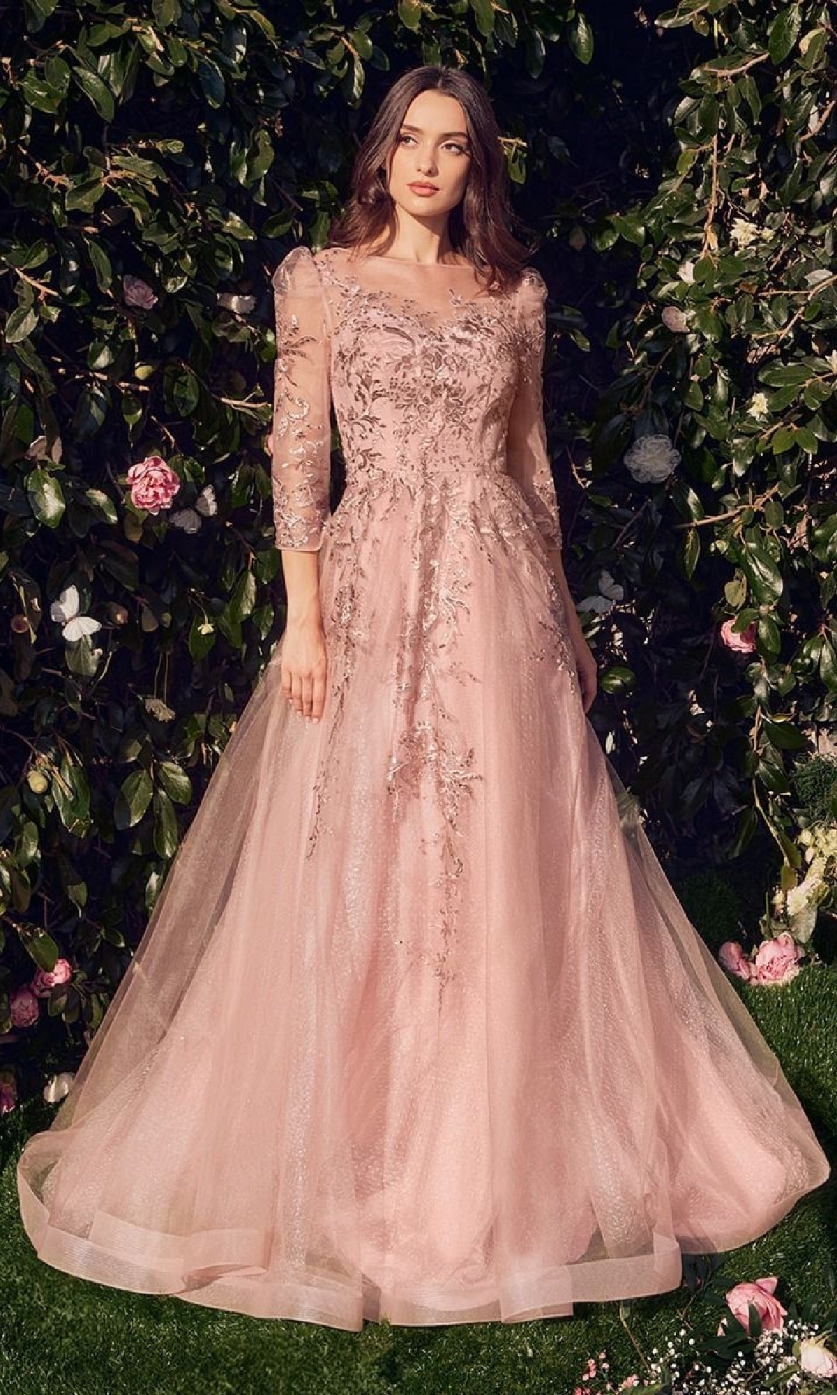 A woman stands outdoors before flowering bushes, wearing the Andrea & Leo A1352 Dusty Rose Pink Formal Dress featuring lace, sheer sleeves, and a long silhouette.