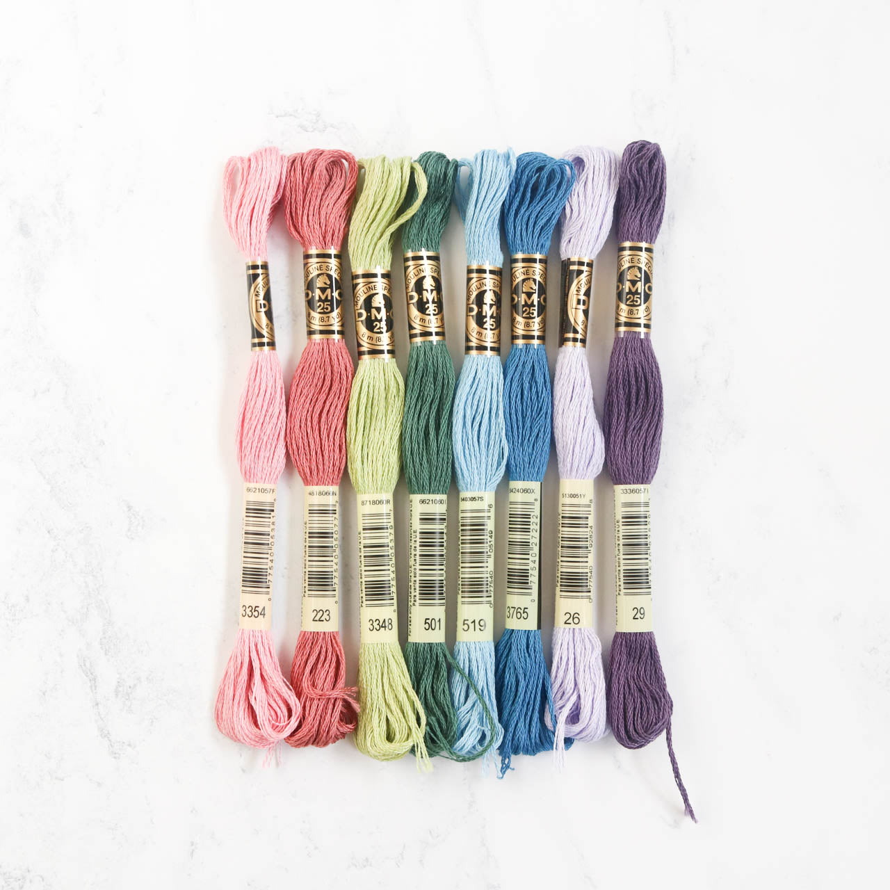 Eight skeins of DMC embroidery floss in pastel rainbow colors lined up vertically on a white marble surface.