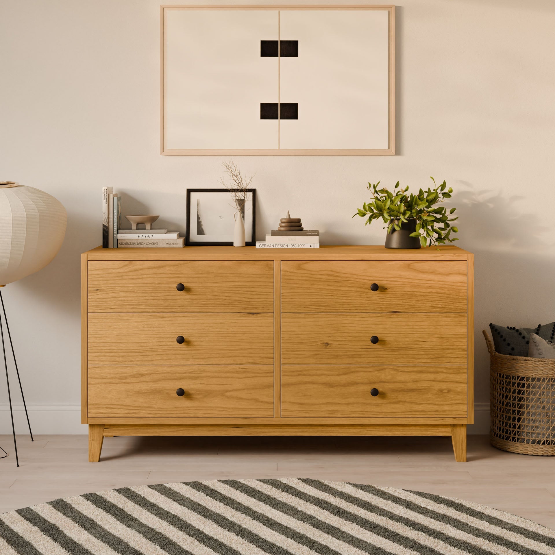 The Maine Made Six Drawer Dresser by Krovel Furniture Co. sits against a light wall with books, framed art, a vase, and a plant on top. Minimalist artwork hangs above; nearby are a basket and floor lamp, with a striped rug below.#wood_cherry